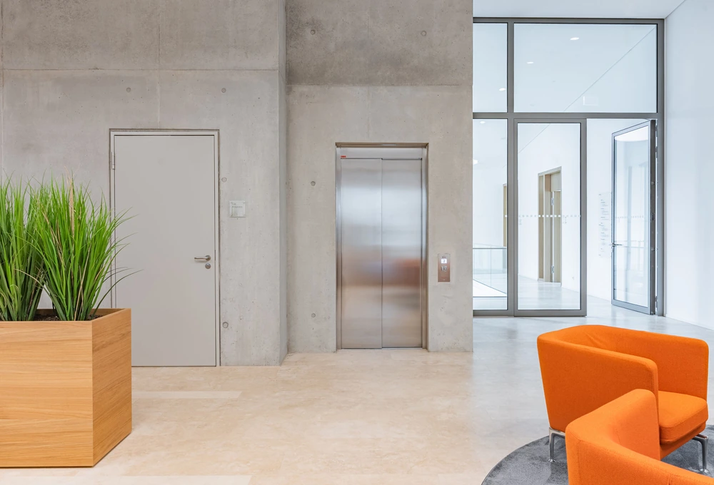 Heller Eingangsbereich mit Aufzug, Glastüren, Betontreppe? No stairs. orange chairs and planter. Need accurate. "Heller Eingangsbereich mit Aufzug, Glastüren, orangefarbenen Sesseln und bepflanztem Holzkübel." Under 125. Note Sessel plural visible 2 chairs maybe. door. Good