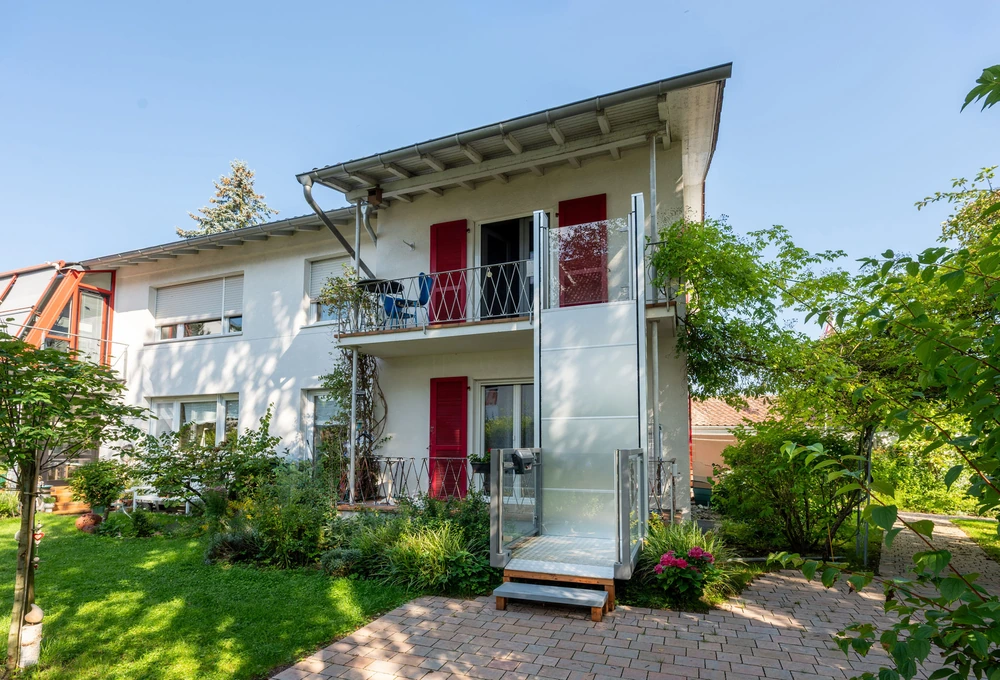 Weißes zweigeschossiges Wohnhaus mit roten Fensterläden, Balkonen, Garten und außen angebrachtem Aufzug
