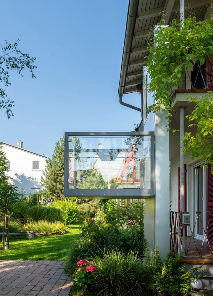 Wohnhaus mit begrüntem Garten, Balkon und vorspringendem gläsernem Anbau an einem sonnigen Tag