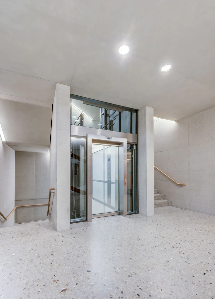 Moderner Aufzug in einem hellen Treppenhaus mit Sichtbetonwänden und Treppen auf beiden Seiten