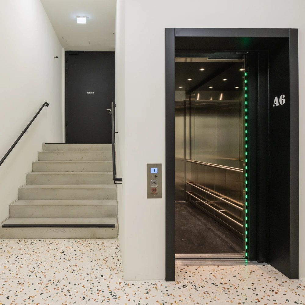 Offener Aufzug neben einer Treppe, mit Anzeige für das erste Obergeschoss