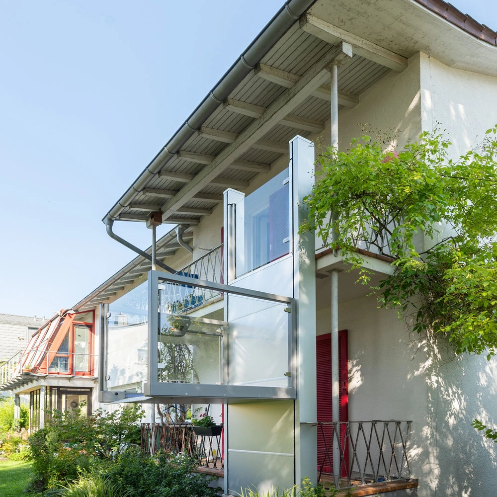 Mehrstöckiges Wohnhaus mit verglasten Balkonen, begrüntem Vorgarten und roten Fensterläden