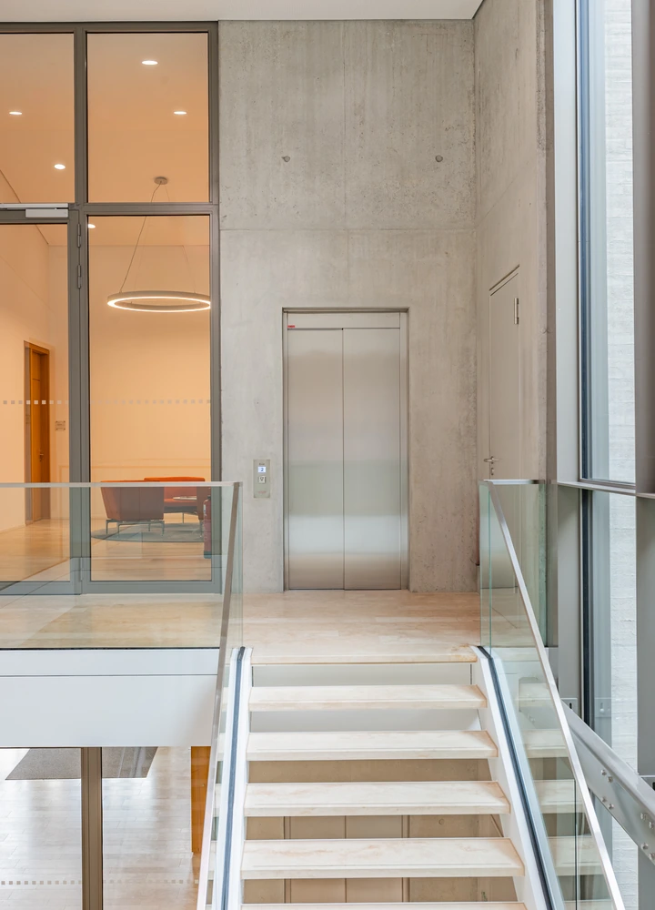 Modernes Gebäudeinterieur mit Treppe, Glasgeländern und Aufzug vor einer Betonwand