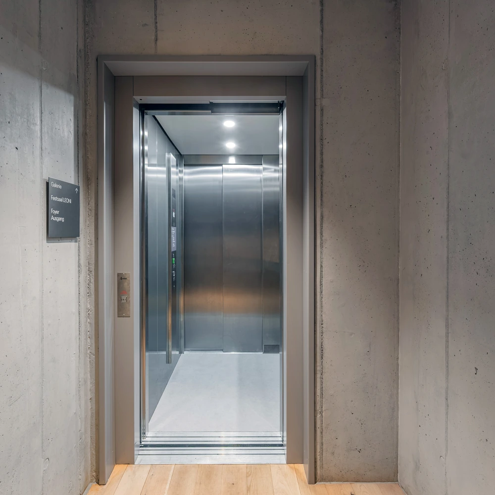 Geöffneter Aufzug in einem Betongebäude, daneben ein Wegweiser zu Galerie, Festsaal, Foyer und Ausgang