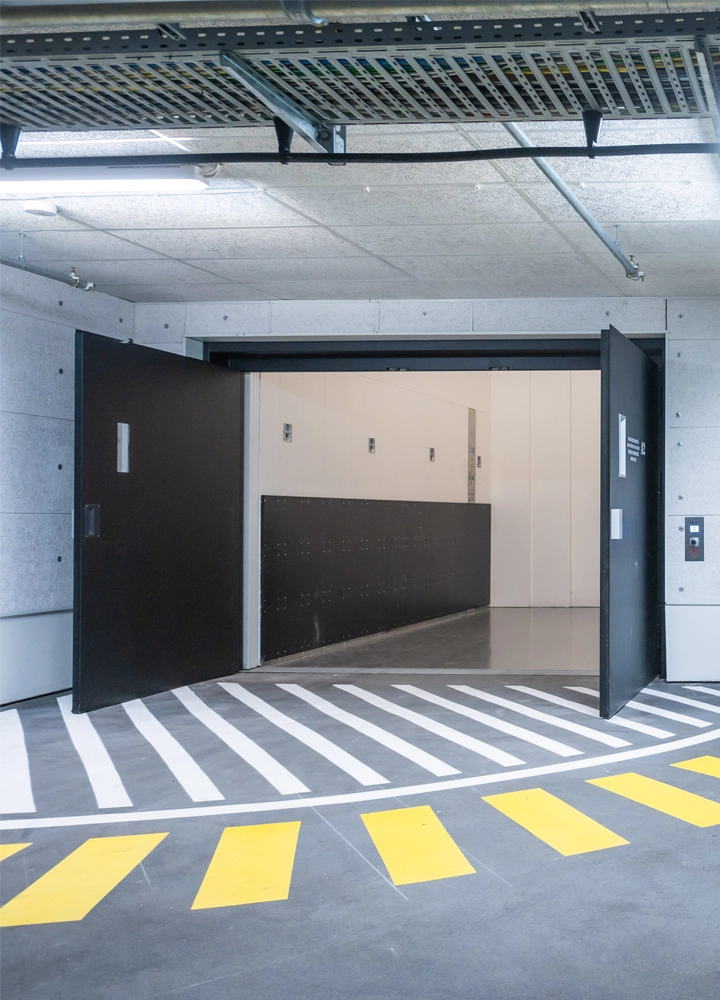 Offener Aufzug in einem Parkhaus mit gelb-weißen Bodenmarkierungen und sichtbaren Kabeltrassen an der Decke