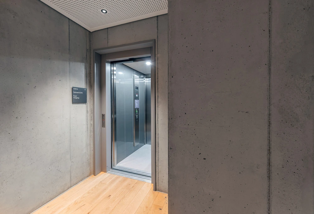 Geöffneter Aufzug in einem Betonflur mit Hinweisschild zu Galerie, FestivalLION, Foyer und Aufgang
