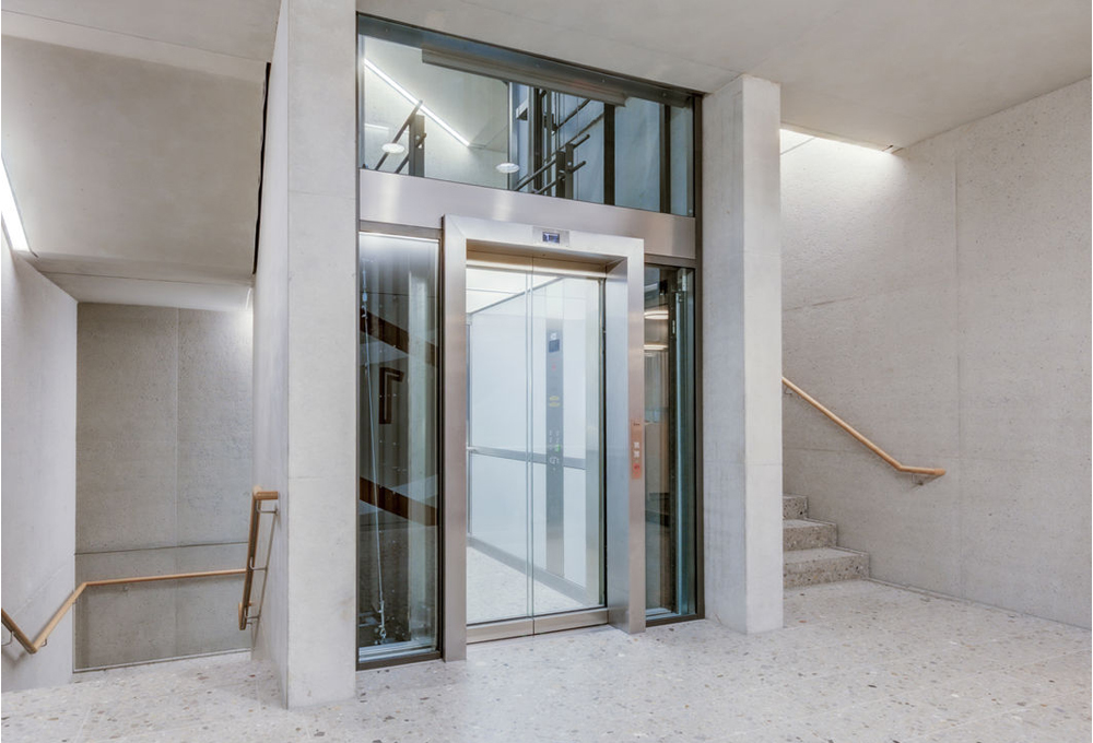 Gläserner Aufzug in einem hellen Treppenhaus mit Betonelementen und Holzhandläufen