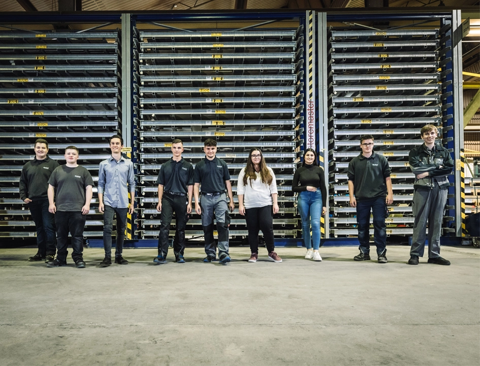 Neun Azubis stehen in einer Lagerhalle vor hohen Regalen mit Metallprofilen