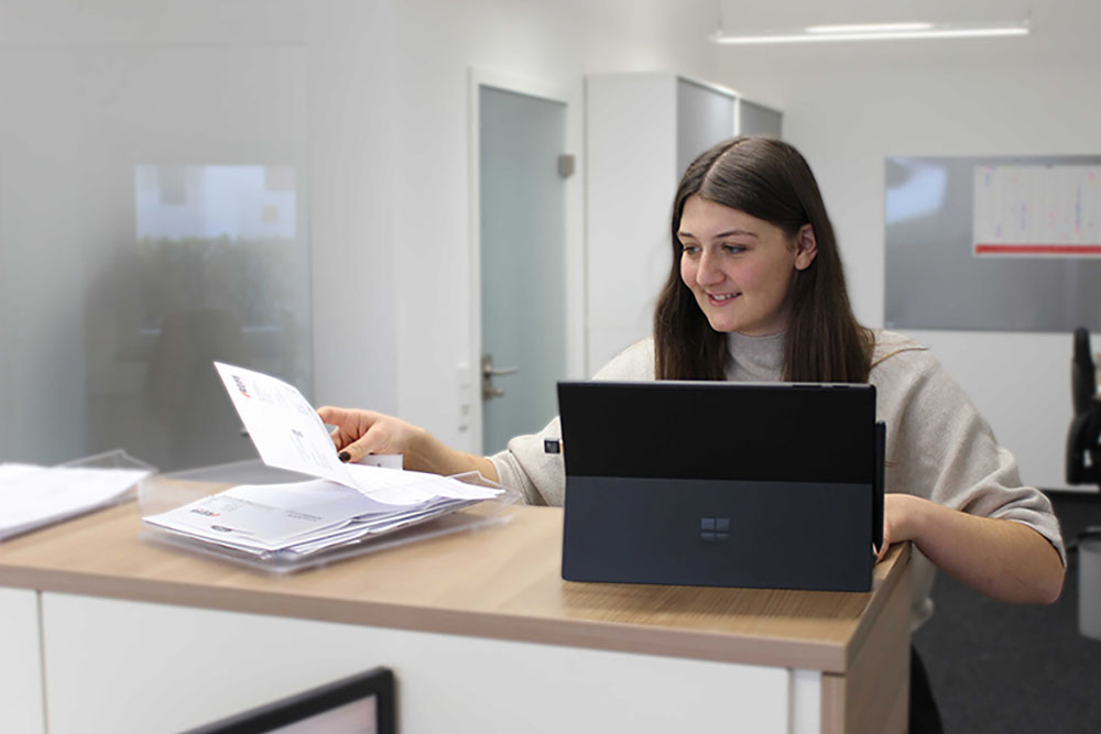 Frau arbeitet an einem Laptop und hält ein Dokument neben einem Stapel Unterlagen in der Hand