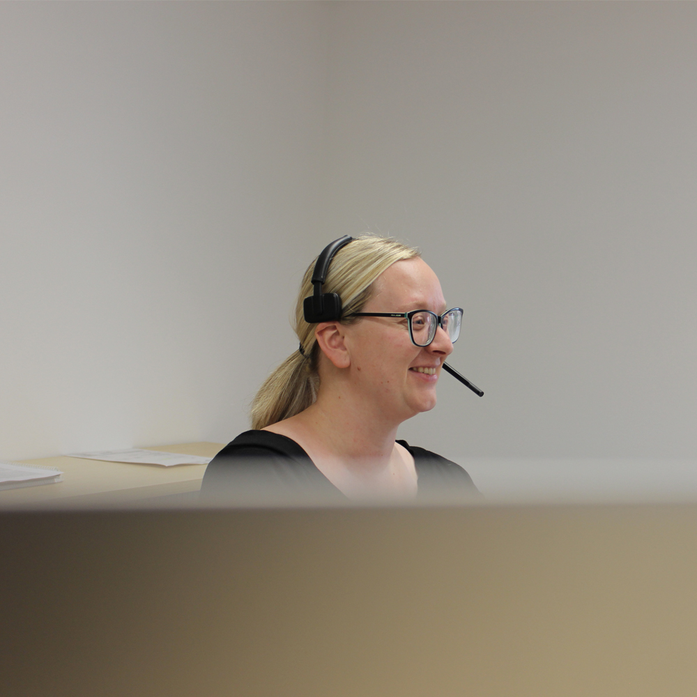 Eine lächelnde Frau mit Brille und Headset sitzt an einem Tisch