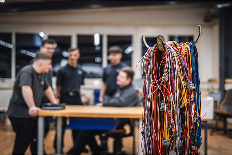 Bündel bunter Kabel im Vordergrund, dahinter mehrere Personen in einer Werkstatt