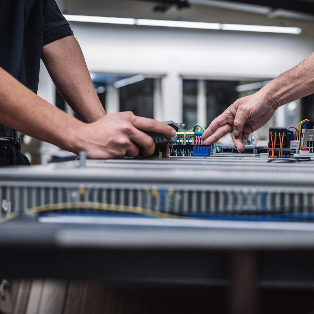 Zwei Personen arbeiten an einer industriellen Schaltanlage; eine Person hält ein Messgerät, während die andere auf die Anschlüsse zeigt
