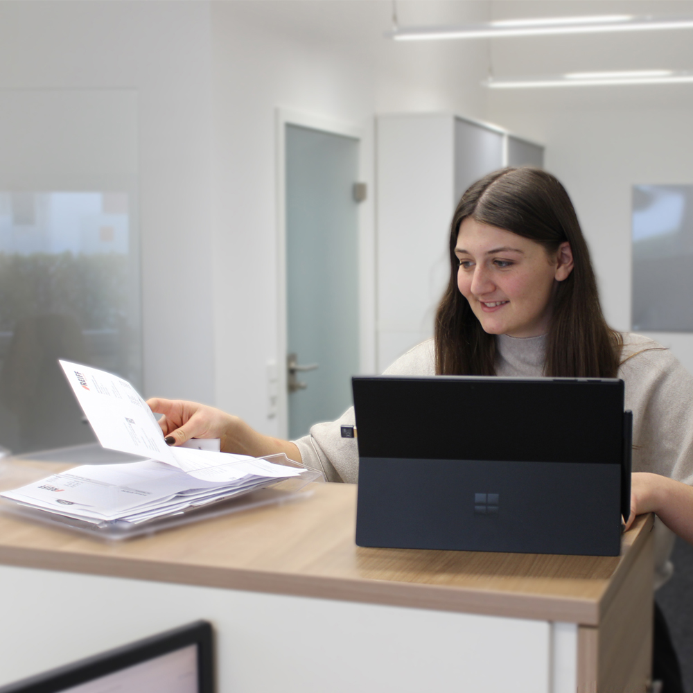 Eine Frau prüft Unterlagen an einem Schreibtisch neben einem geöffneten Laptop