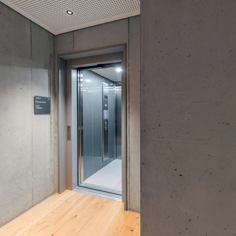 Geöffneter Aufzug in einem Betonflur; ein Schild weist zur Galerie, zum Festsaal LEON, Foyer und Ausgang