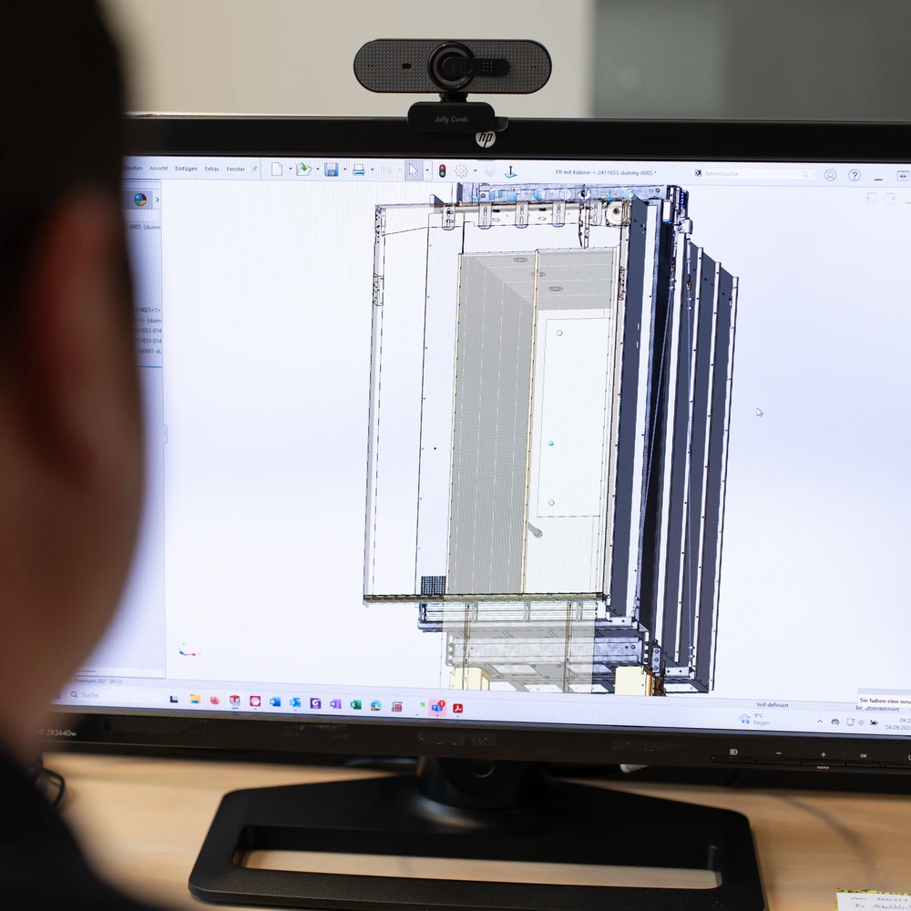 Auf einem Monitor ist eine technische CAD-Zeichnung eines rechteckigen Bauteils zu sehen; darüber befindet sich eine Webcam