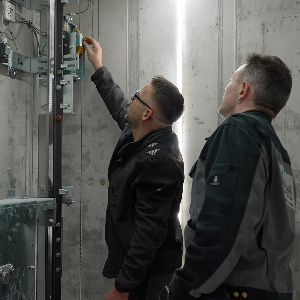 Zwei Techniker prüfen die Komponenten einer Aufzugsanlage; einer betätigt ein Bauteil am Führungssystem