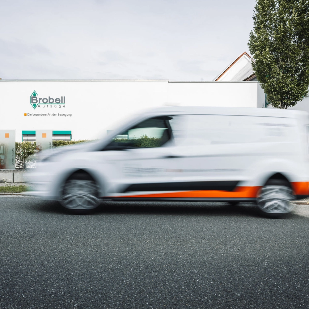 Vor einem Gebäude mit dem Logo „Brobeil Aufzüge“ fährt ein weißer, orange-grün gestalteter Transporter vorbei